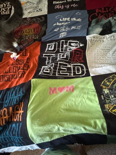 Close-up of a customer's premium t-shirt quilt showing various patches, including one with 'MOM' in pink, with a cat on the edge.