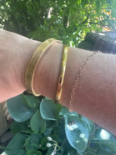 Customer wearing the gold Marbella Cuff Bracelet stacked with two other bracelets outdoors against a leafy background.