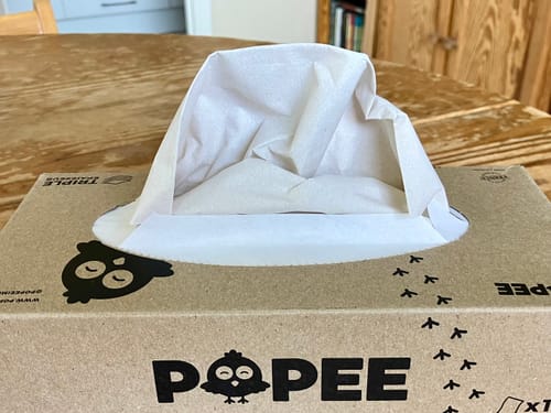 Un client montre la boîte de mouchoirs TRIPLE Épaisseur sur une table en bois, avec un mouchoir qui en sort.