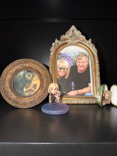 Customer's custom 3D figurine of a woman and dog, displayed on a dark shelf next to a matching photo.