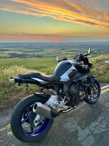 Customer's black Yamaha MT10 with blue wheels parked on a scenic road overlooking a valley at sunset.