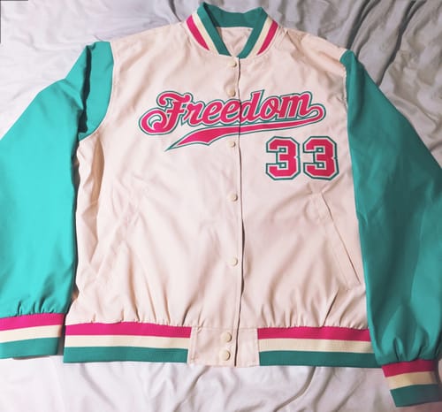 Customer's custom varsity jacket laid flat, showing the front with 'Freedom 33' stitched in pink and aqua on a cream body.