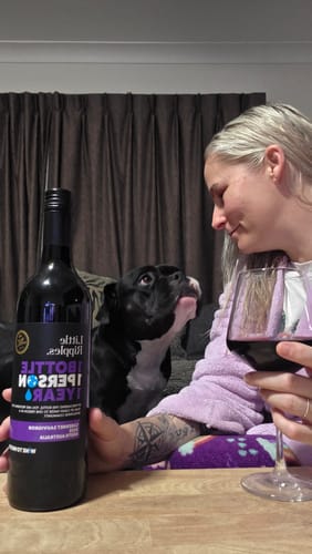 Reviewer at a table with a bottle and glass of red wine from the Fan Favourites pack, smiling at their dog.
