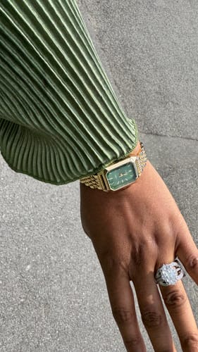 A close-up of a reviewer wearing the gold CLAIRE DE MAINS watch with a green rectangular face, paired with a pleated green sleeve.