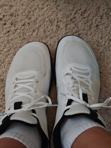 A customer's feet from a top-down perspective, wearing the white CUURT Muuv Wide Toe Box Court Shoes on a carpet.