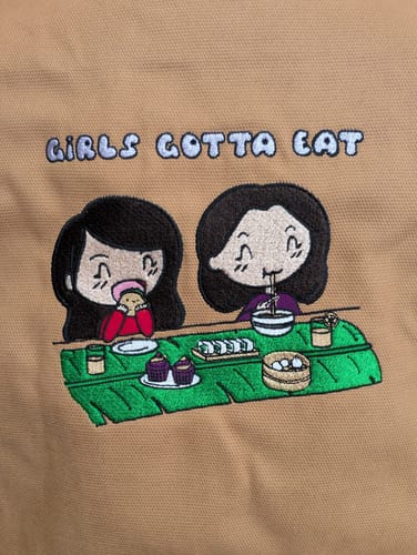 Close-up on a customer's tan canvas tote showing the detailed custom embroidery of two cartoon girls eating.