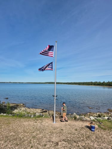 Customer installing the tall Commercial Flagpole on a shore, with two flags flying from the top against a clear blue sky.