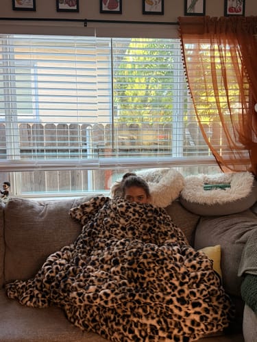 Customer snuggled on a couch under the plush Hugs Golden Leopard Blanket, peeking over the top.