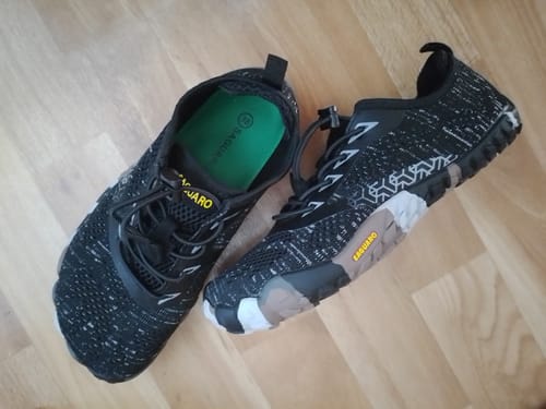 Ein Kunde zeigt seine schwarzen Smart II Barfußschuhe mit gemustertem Design auf einem Holzboden.