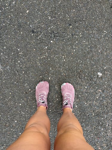 A reviewer's first-person view of their pink Smart II Barefoot Shoes on an asphalt road.