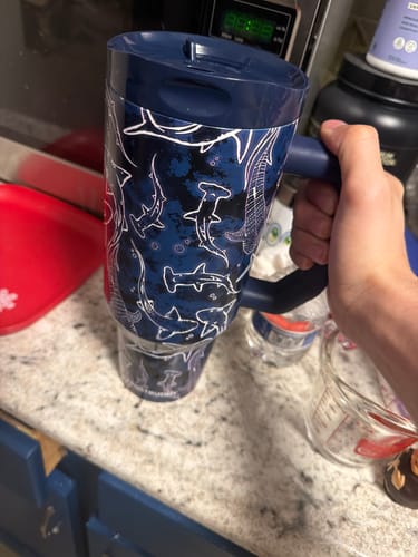 Customer holding their Thicc Buddy 40oz mug with the dark blue Midnight Reef shark pattern in a kitchen.