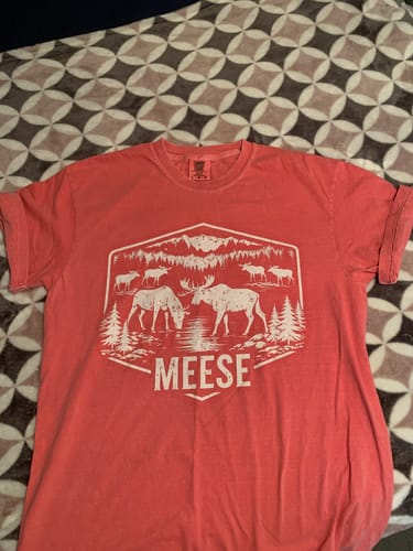 Customer's coral Meese T-shirt with a white moose graphic laid flat on a patterned surface.
