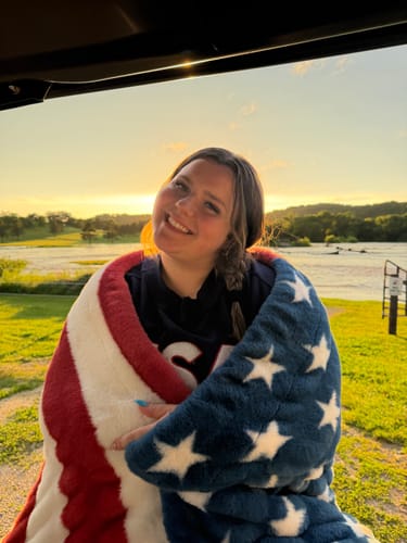 Customer smiling while wrapped in a plush Hugs Flag Blanket outdoors at sunset.