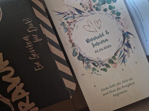 Kunde zeigt den personalisierten Inhalt für die Holzkiste mit Namen, Datum und einem Blumenmotiv neben der Verpackung.