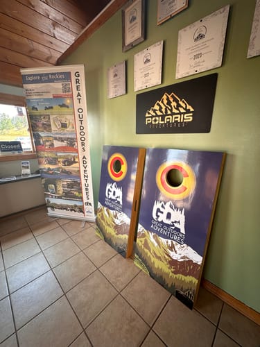Customer's two Custom Cornhole Board Wraps with a mountain and 'Great Outdoors Adventures' logo, standing upright indoors.