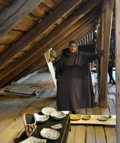 Reviewer in a black, hooded drawstring dress standing in an attic setting during a ceramic exhibition.