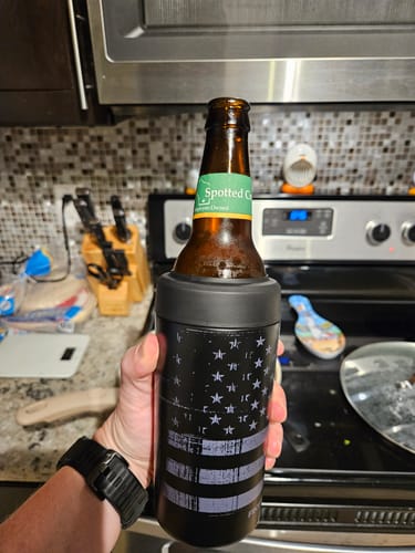 Reviewer's hand holding a gunmetal Universal Buddy can cooler with a glass bottle inside, in a kitchen setting.