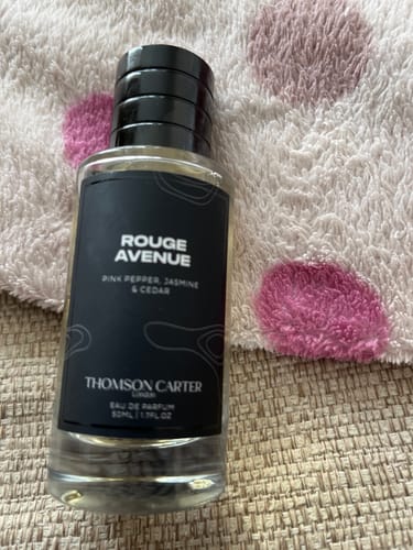 Customer's glass bottle of Rouge Avenue Eau de Parfum featuring a black label, resting on a soft pink polka dot surface.