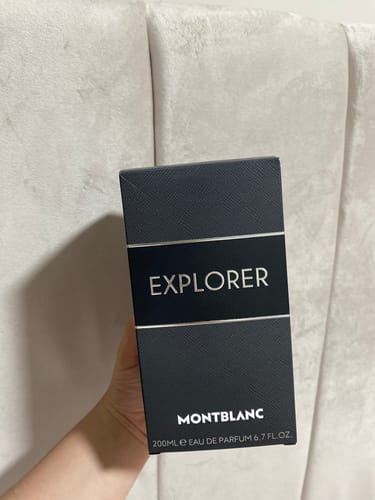Customer's hand holding the black Montblanc Explorer 200ml perfume box in front of a white padded headboard.