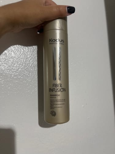Customer's hand holding a gold bottle of Kadus Fiber Infusion Shampoo against a white wall.