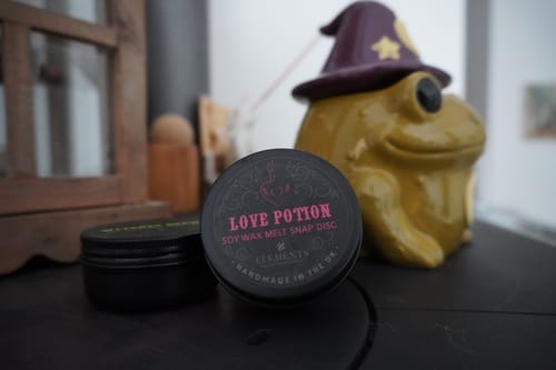 Kundenfoto des Love Potion Duftwachses in einer schwarzen Dose neben einer grünen Froschfigur mit Hexenhut.