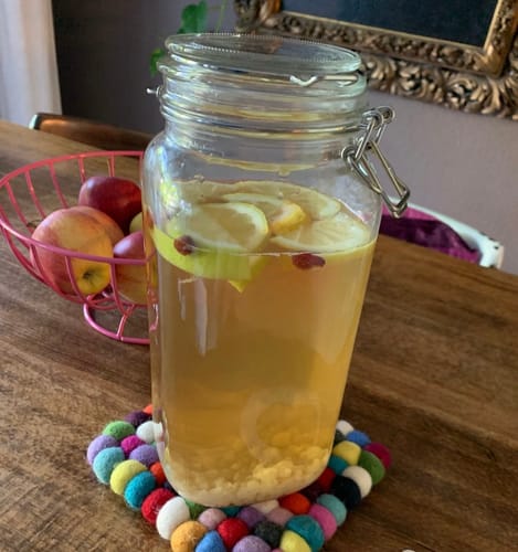 Kundenfoto von angesetztem Wasserkefir in einem Glasgefäß mit Zitronenscheiben und Früchten auf einem Tisch.