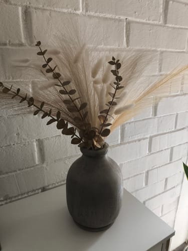 Customer's Florero Madera redondo de madera oscura sobre una superficie blanca frente a una pared de ladrillo blanco.