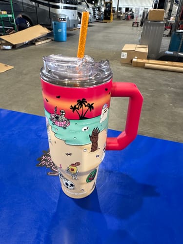 Customer's colorful Frost Buddy insulated tumbler with beach-themed graphic, featuring an orange straw on a blue table.