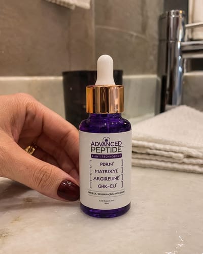 Customer's hand holding the purple glass bottle of Advanced Peptide serum with a white dropper cap in a bathroom.