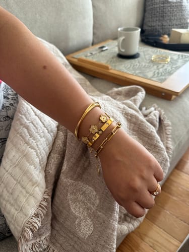 Customer's wrist wearing multiple Gold Clover Bangles styled with other metallic bracelets on a couch.