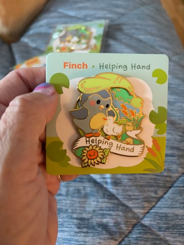 Customer's hand holding the Helping Hand Pin, featuring a cute bird design on its display card.