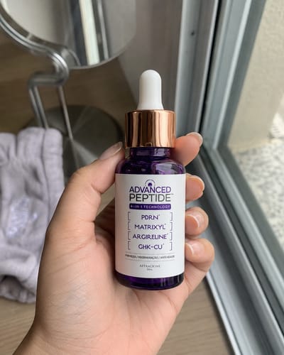Customer's hand holding a purple glass bottle of Advanced Peptide serum with a white dropper cap.