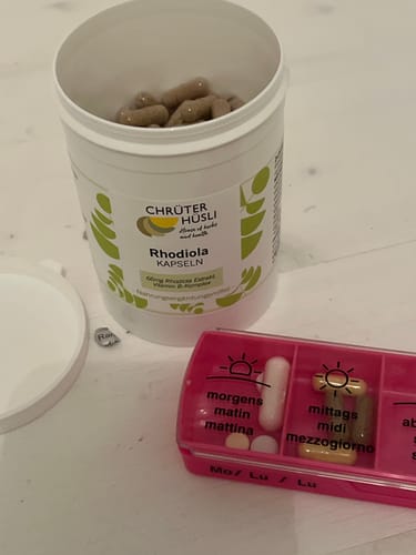 Customer's Rhodiola Kapseln bottle with capsules visible inside and a pink weekly pill organizer on a surface.