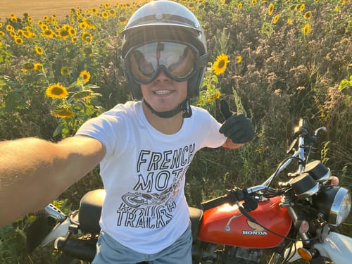 Client en tee-shirt Collection Mc Queen, portant un casque et des gants, posant avec une moto rouge dans un champ.