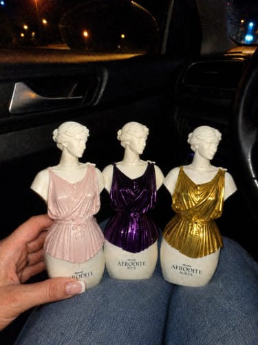 Customer's Kit Afrodite set of three statue-shaped perfume bottles in pink, purple, and gold, held in a car.