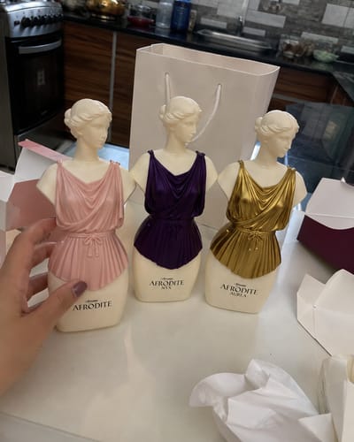 Customer's Kit Afrodite perfume bottles in pink, purple, and gold, shaped like statues, on a counter.