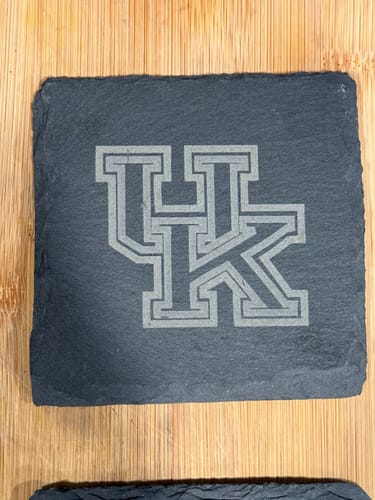 Customer's square, dark slate coaster engraved with a University of Kentucky UK logo on a wooden surface.