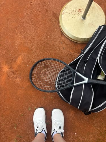 Customer's white CUURT Muuv court shoes worn on a clay tennis court with a racquet and sports bag nearby.