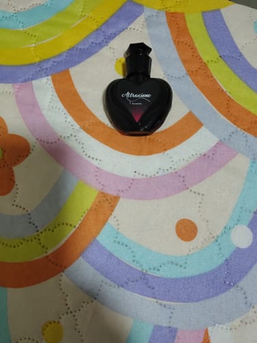 Reviewer's Perfume Attracione Sensual, a small black heart-shaped glass bottle resting on a colorful patterned surface.