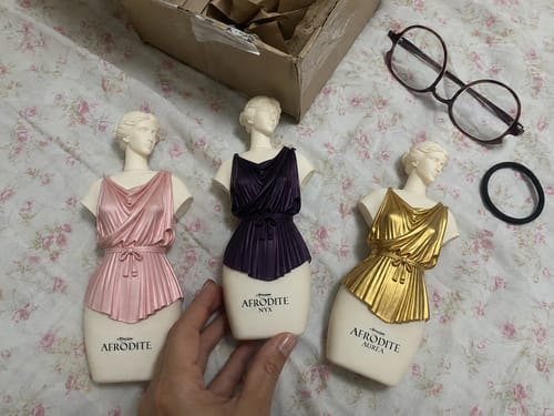 Reviewer's three Kit Afrodite fragrance bottles shaped like elegant torsos in pink, purple, and gold on floral fabric.