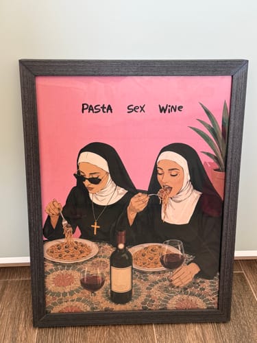 Reviewer's Black Oak Gallery Frame containing an art print of two nuns eating pasta and drinking wine on a pink background.