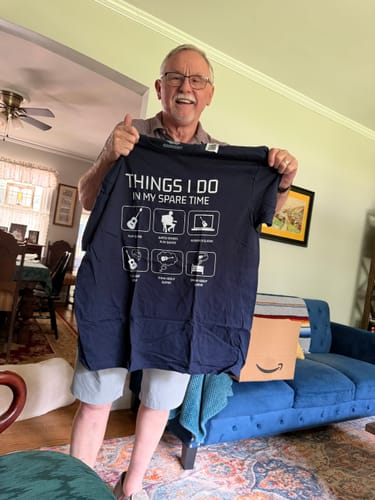 Customer's navy All I Do is Guitar T-Shirt with graphic print, held up by a smiling person in a living room.