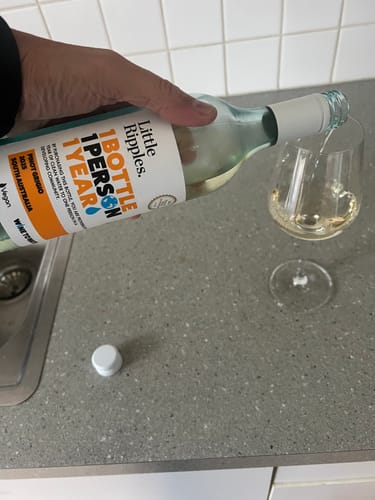 Customer's hand pouring clear Little Ripples Pinot Grigio from a bottle into a glass on a kitchen counter.