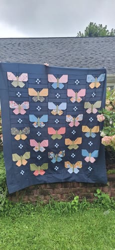 Customer's Flutter Lella Boutique quilt displayed outdoors, featuring colorful butterfly patterns on dark blue fabric.