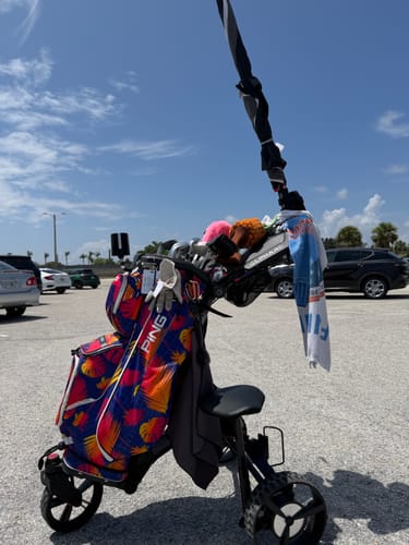 Customer's Cybercart golf caddy with a colorful PING bag and umbrella, parked on a paved lot.