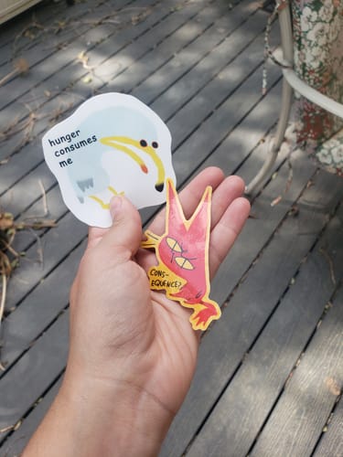 Customer's hand holding two custom vinyl stickers, one white and one yellow-bordered red cat, over a wooden deck.