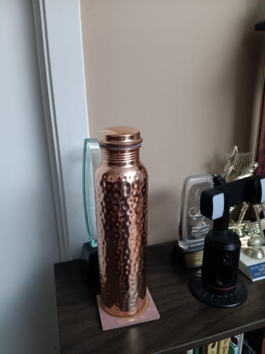 Customer's hand-hammered copper water bottle from Certified Ayurveda, standing on a wooden surface.