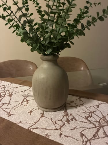 Customer's Florero Madera redondo de madera gastada con hojas verdes, colocado sobre un camino de mesa decorativo.