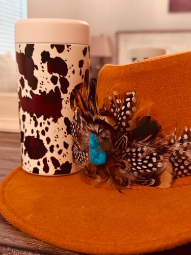 Customer's cow-patterned Universal Buddy can cooler sitting next to an orange felt hat decorated with feathers
