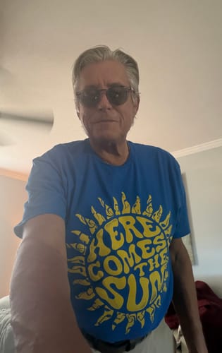Reviewer wearing a blue Here Comes The Sun T-shirt with a yellow sun graphic while posing indoors.